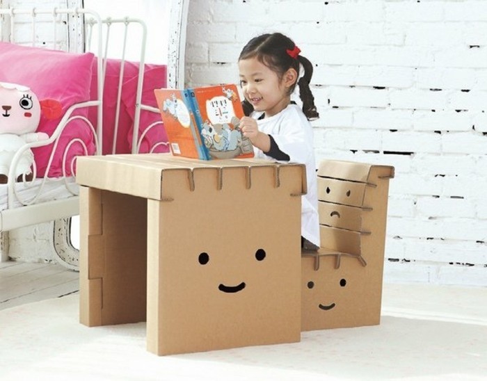 möbel aus karton für das kinderzimmer. ein kleines mädchen sitzt am schreibtisch aus karton und auf einem lustigen kartonstuhl, auf möbel zeichnen, emoji, gesicht zeichnen, kinderbuch lesen