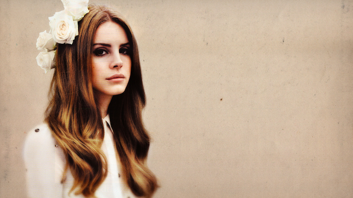 Lana Del Rey mit weißen Rosen im Haar, hellbraune wellige Haare mit Mittelscheitel, Smokey Eyes und matter Lippenstift 