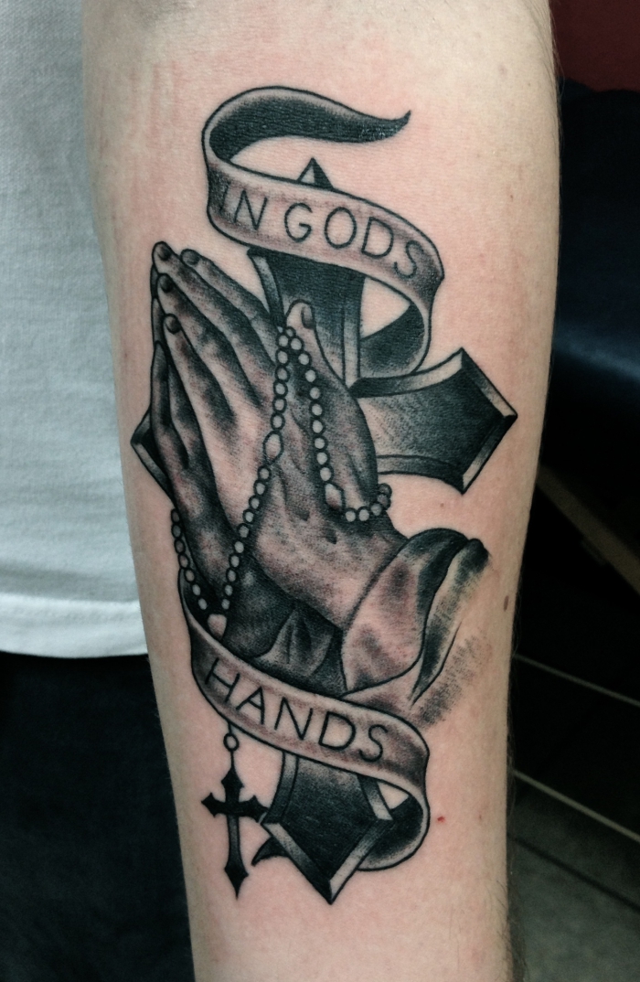 rosenkranz tattoo in kombination mit bettenden händen und schriftzug, schwarz graue tätowierung am unterarm