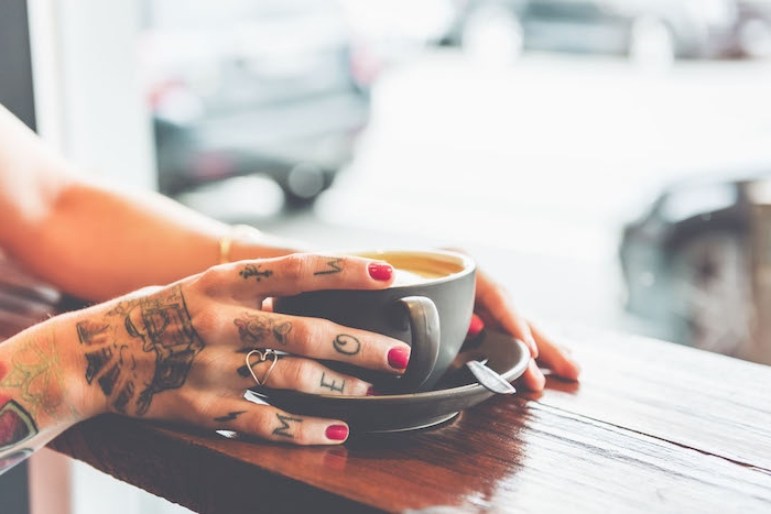 Farbige Tattoos an der Hand, roter Nagellack, herzförmiger silberner Ring, Tasse Kaffee 
