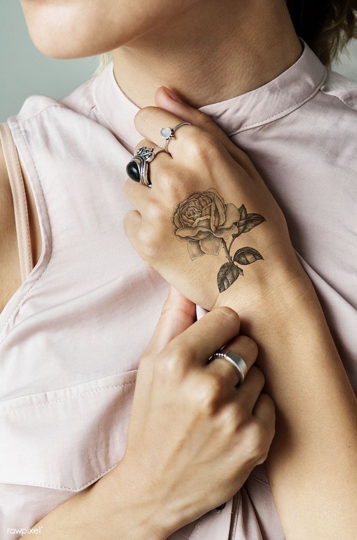 Zartes Tattoo Rose an der Hand, silberne Ringe am Mittelfinger und Zeigefinger, Top in Hellrosa 