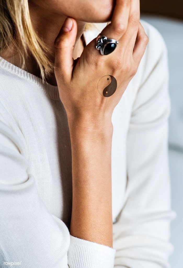 Yin und Yang Tattoo an der Hand, silberner Ring mit schwarzem Stein am Mittelfinger, weiße Bluse, blonde Haare 