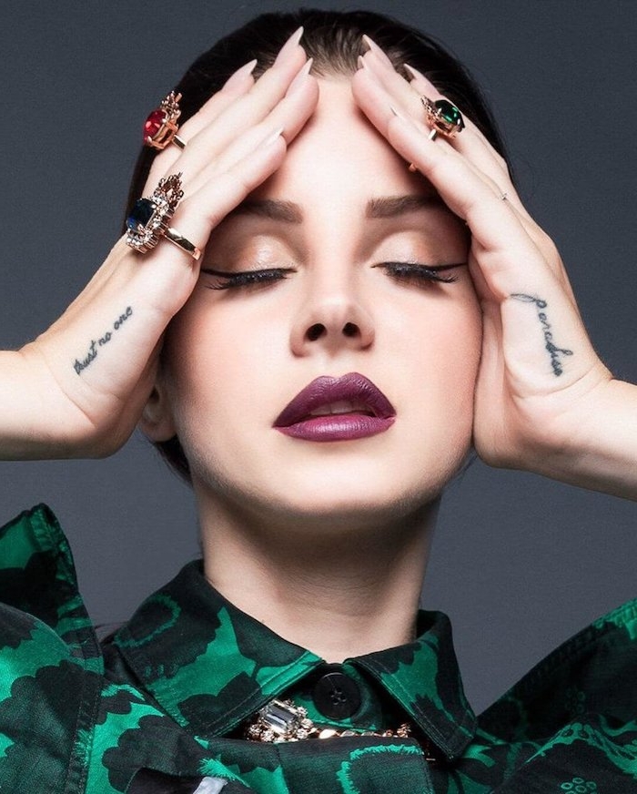 Lana Del Rey Hand Tattoos, Ringe mit großen Steinen, lila Lippenstift und schwarzer Eyeliner, Hemd in Grün Nuancen 