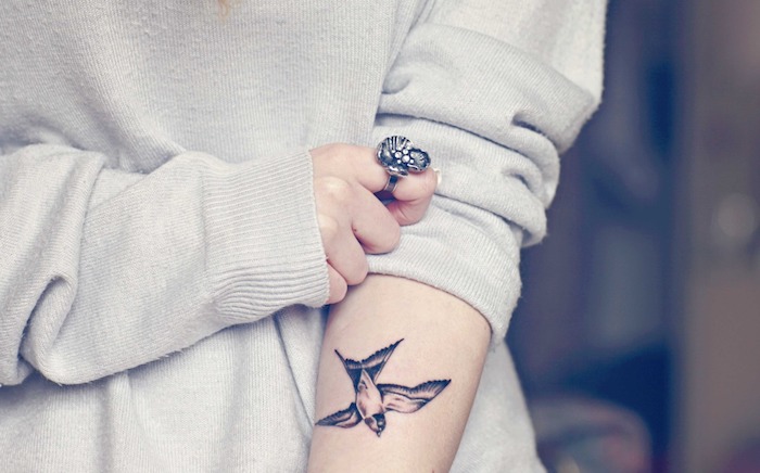 Ideen für Vögel Tattoos, kleine Schwalbe am Unterarm, Symbol für Freiheit, weißer Pullover, massiver Ring am Mittelfinger 