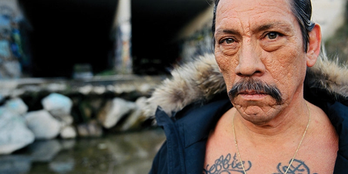 Danny Trejo mit zwei Tattos, eine schwarze Jacke, großer Schnurrbart, ein Wasserspiel 