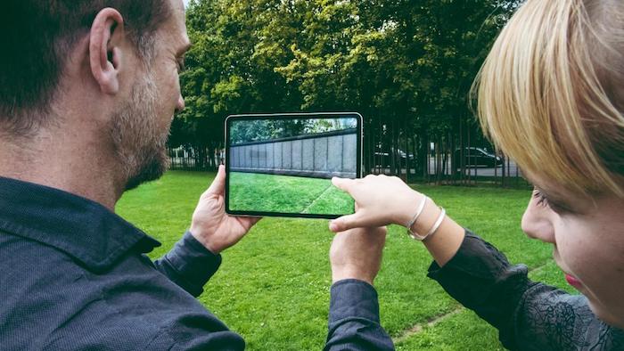 garten mit grünen bäumen und der berliner mauer, ein man und eine frau mit hand mit armband, ein tablet mit der neuen app mauAR, die die ehemalige berliner mauer zeigt