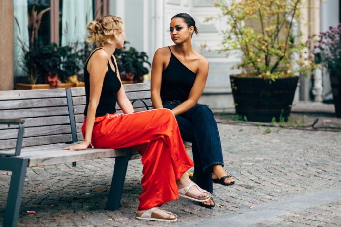 schwedische mode für damen, zwei freundinnen sitzen auf einer bank und sprechen, rote hose, dunkler jumpsuit