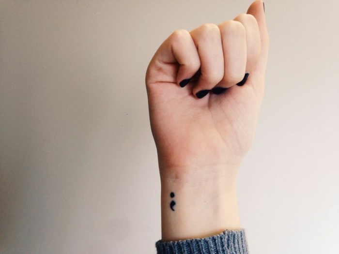 Gebogene Hand mit dunklem Nagellack mit einem Semicolon Tattoo, grauer Pullover, semikolon bedeutung