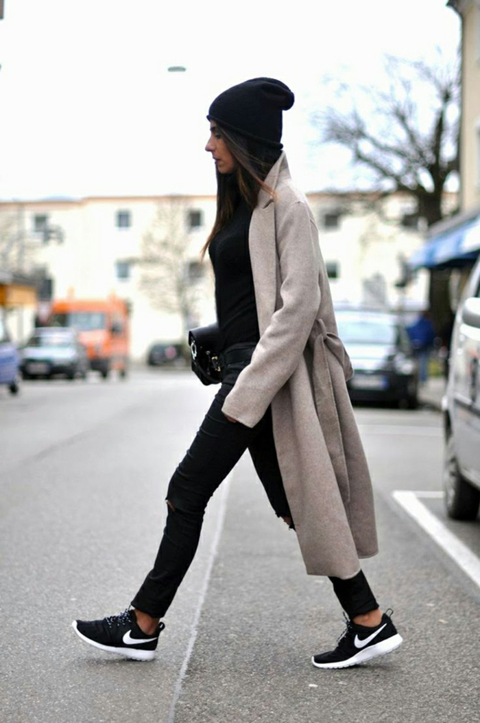 Bequem angezogene Frau in Jeans und Sneakers in Schwarz, mit Mütze und schwarzer Tasche