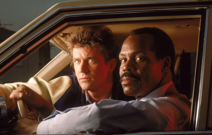 die schauspieler danny glover und mel gibson, ein mann mit langem haar, eine szene aus dem film lethal weapon, zwei männer in einem auto 