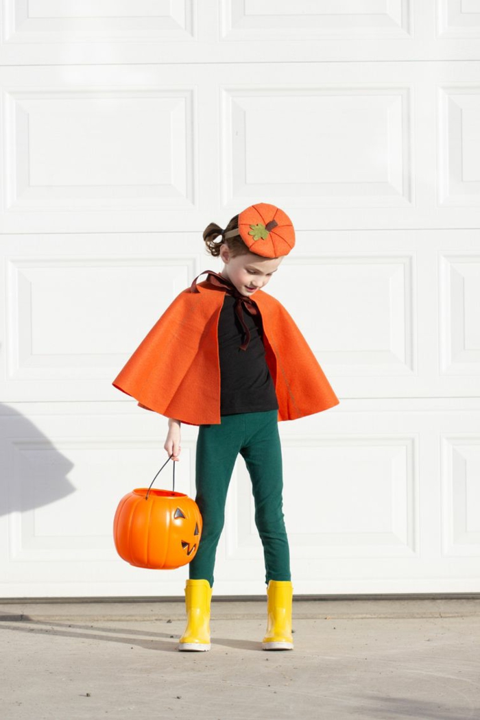 Faschingskostüme für Kinder, Kürbis Kostüm mit Umhang und Hut in orange mit grünen Hosen und gelben Schuhen, Mädchen hält ein Kürbiskopf