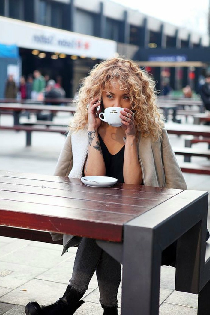 Frau mit blonden Haaren trinkt Kaffee draußen, Tattoo auf der Hand, Kurzhaarfrisuren mit Locken, beige Jacke