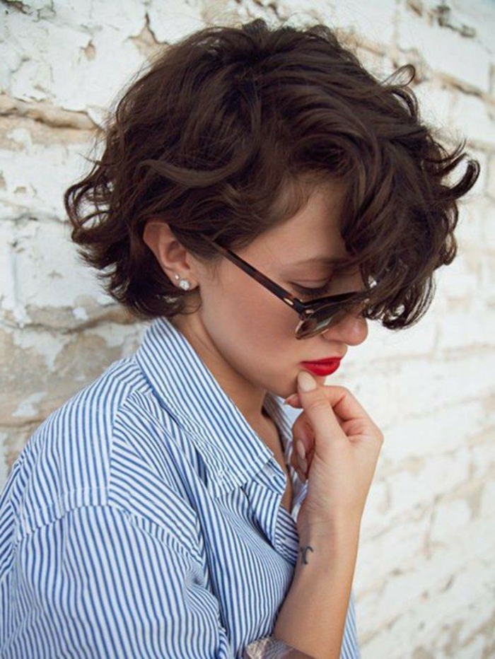 Kurzhaarfrisuren Damen Bilder, braune Haare mit Locken, runde Sonnenbrillen in schwarz, roter Lippenstift, weiß blaue Bluse gestrift