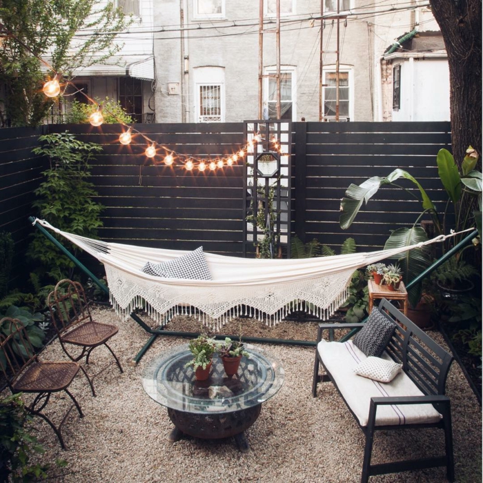 cozy Einrichtung Garten mit weißer Hängematte, Sichschutz aus Holz in braun, aerufgehängte Lichtketten, Boden mit Zierkies, runder Glastisch, gartendeko ideen