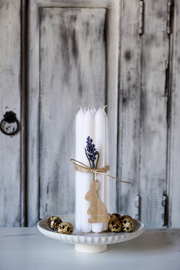 große weiße Kerze auf einem Kuchenteller, umwickelt mit einem Band mit einem hängenden Hasen aus Holz. Osterdeko Holz