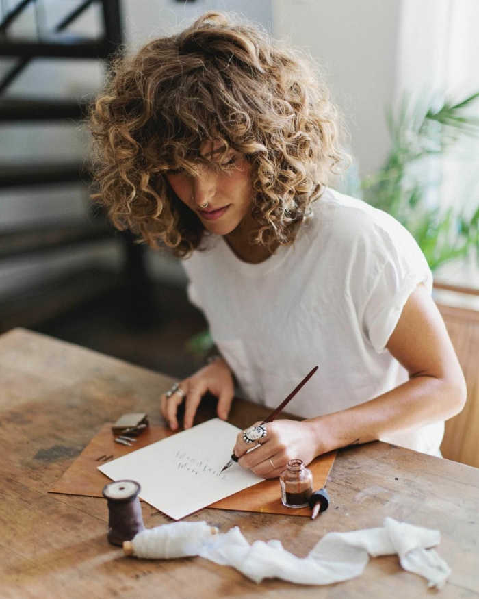 Frau im weißen T-Shirt schreibt eine Karte, Haarschnitt für lockiges Haar, moderne Kurzhaarfrisuren, Federhalter und Tinte