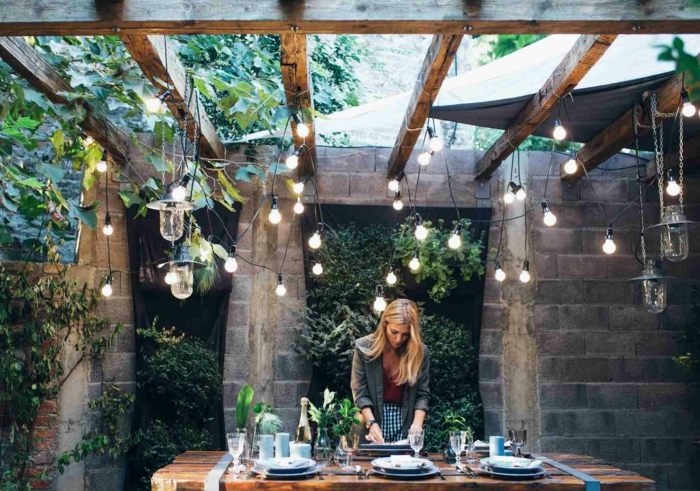 Blonde Frau deckt Tisch in einem Garten im rustikalen Stil. hängende Lichterketten, Gartendeko Ideen, grüne Pflanzen Dekoration