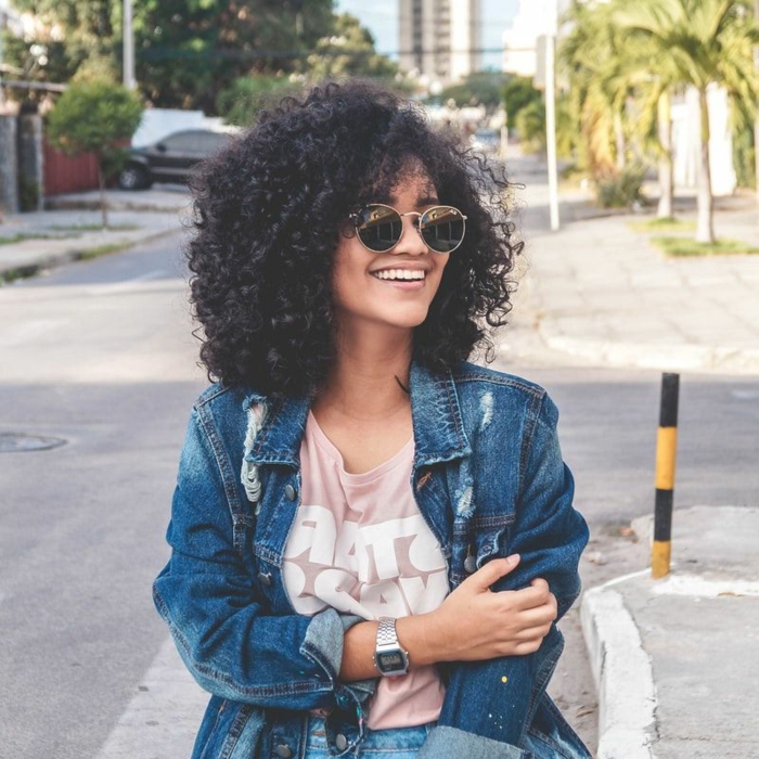 lächelnde junge Frau, gekleidet in Jeansjacke und pinkes T-Shirt, Naturlocken kurz schneiden, runde Sonnenbrillen