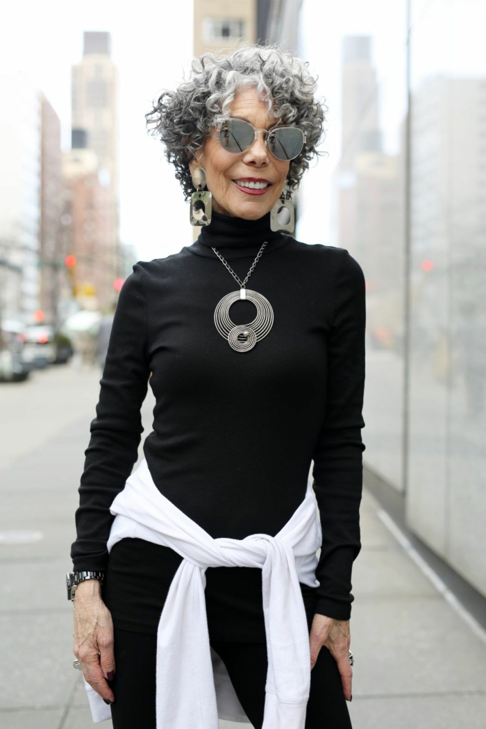 Elegante Dame mit grauen lockigen Haaren, schwarzes Outfit mit Rollkragenpullover, schicke Kurzhaarfrisuren, eindrucksvolle Halskette