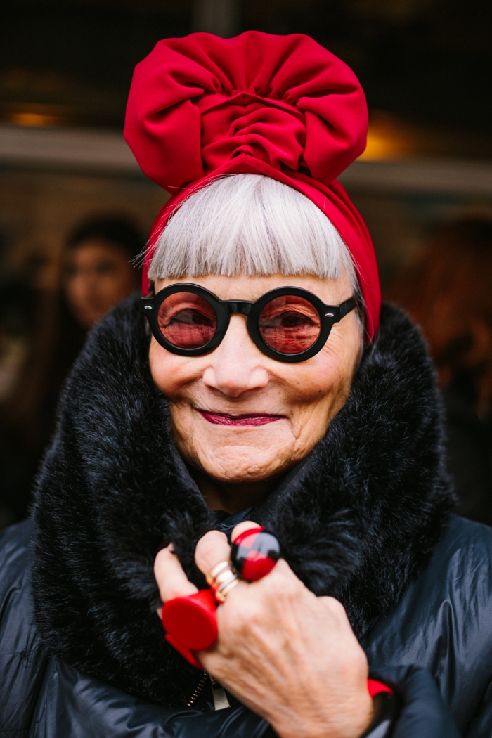 eindrucksvoll gekleidete ältere Dame, runde rote Sonnenbrillen und große Ringe. weiße Haare, micro Pony, effektvoller Hut