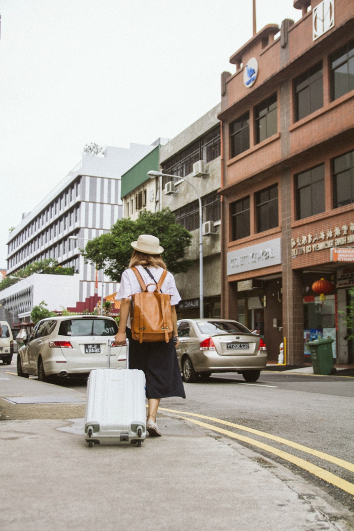Frau mit einem schwarzen Rock und weißes T-Shirt, zieht einen kleinen Handgepäck Koffer, brauner Rucksack aus Leder, Reisegepäck wählen