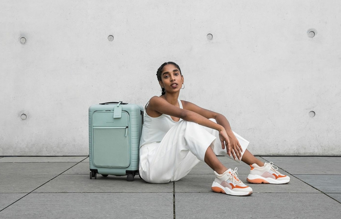 Frau im monochromen weißen Outfit und weiß orange Sneakers, kleiner blauer Handgepäck, Reisegepäck wählen