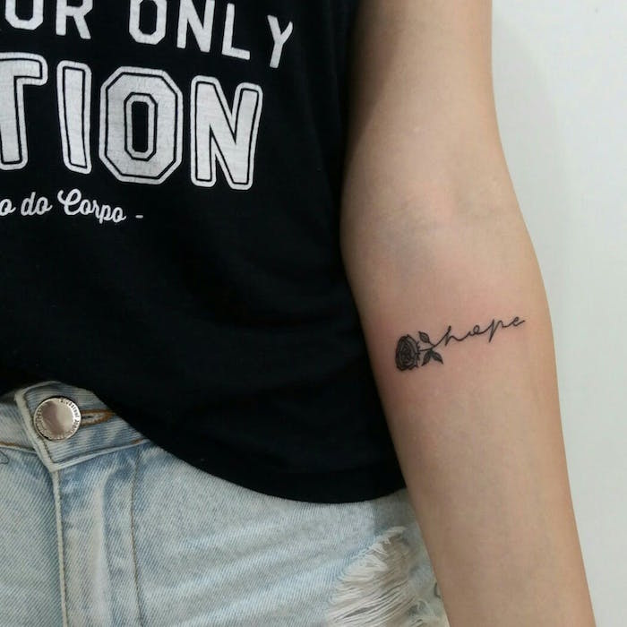 Schönes Tattoo am Unterarm, kleine Rose und das Wort Hope, schwarzes Top mit weißer Aufschrift, Jeans mit hoher Taille 