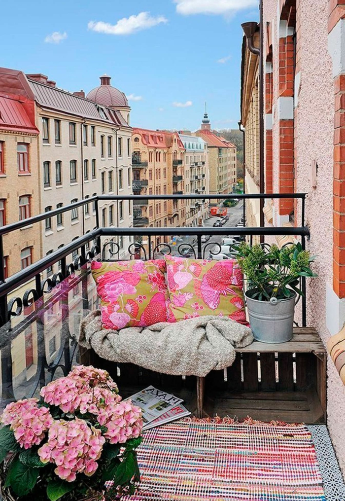 Möbel für kleinen Balkon, Sitzbereich aus alten Kisten, zwei pinke Kissen und bunter Teppich, Blume mit rosa Blüten