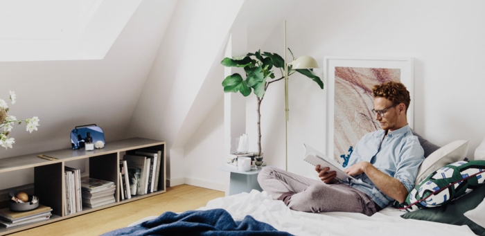 Mann liest eine Buch im Bett, kleine offene Kommode, große grüne Pflanze Tylko Möbel