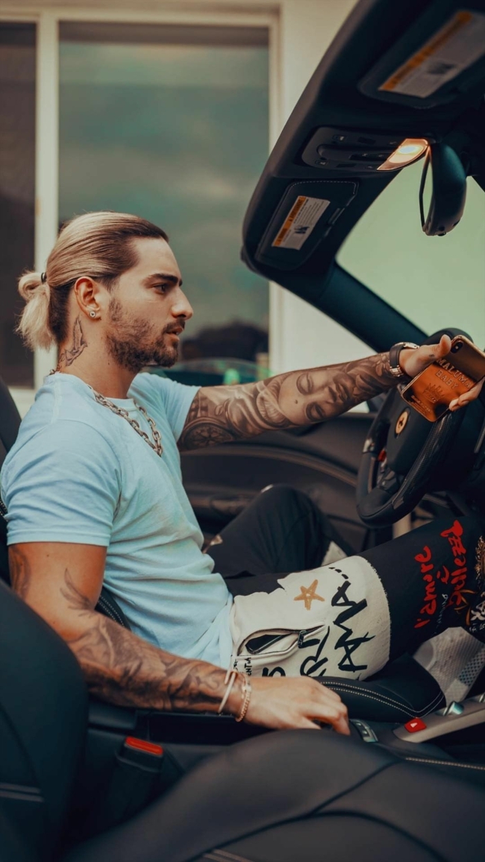 blonder Mann sitzt in einem Auto, Mittellange Haare Frisur 2020, Tattoos auf beide Hände, blaues T-Shirt und bunte Hose