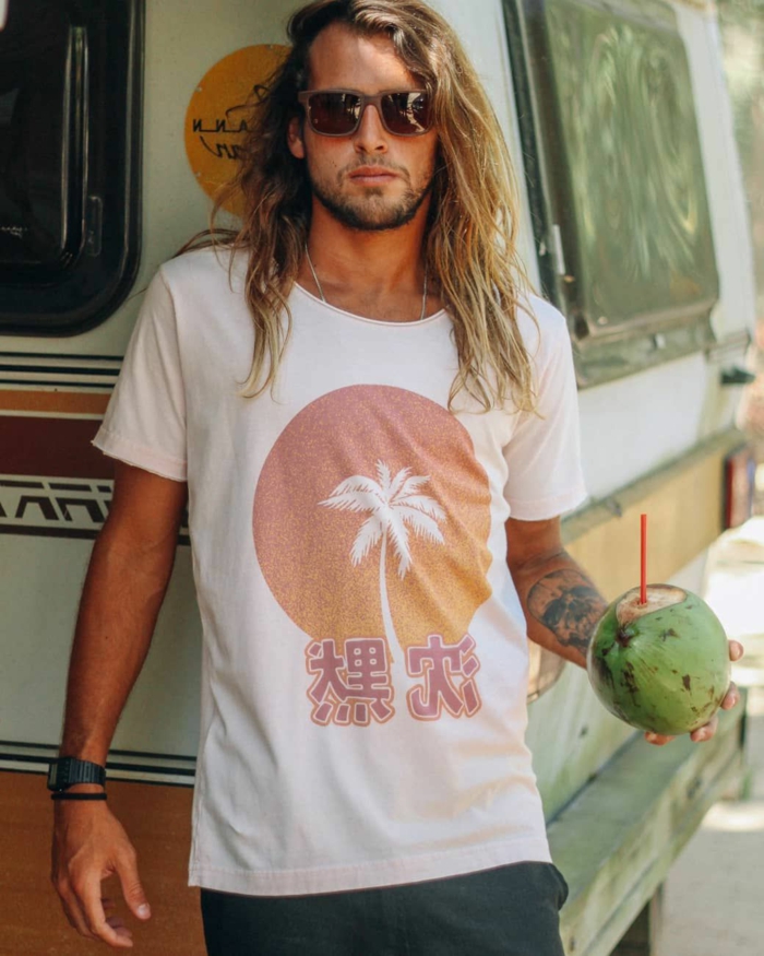 Männerfrisuren lang, Mann mit blonden Haaren, angezogen im weißen T-Shirt mit Abdruck, hält eine Kokosnuss in der Hand