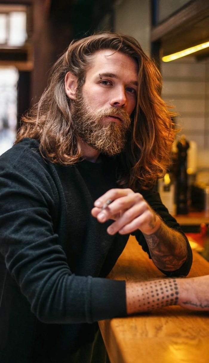 Mittellange Haare Frisur mit buschigem Bart, Tattoos auf den Armen, Mann angezogen in schwarze Bluse, dunkelblonde Haare