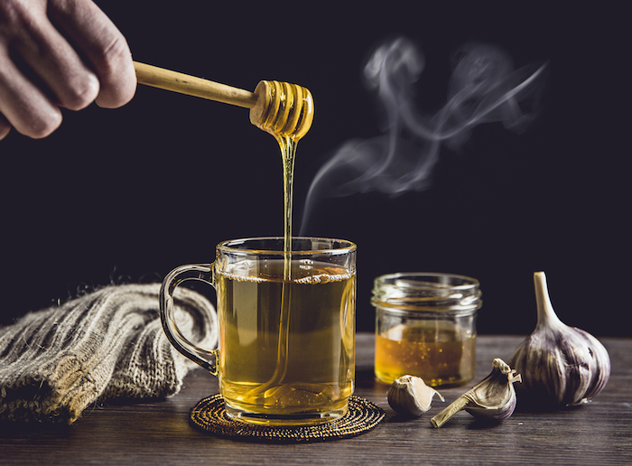 man hand holding wooden honey dipper, honey spoon on top of glass of tea/ medicine and dripping honey in hot tea. knitted socks, small jar of honey, garlic on wooden table against black background.