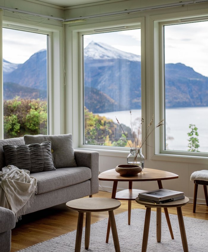 kleine und große tische aus holz graues sofa mit kissen skandinavisches wohnzimmer einrichten große fenster mit aussicht zum see und bergen