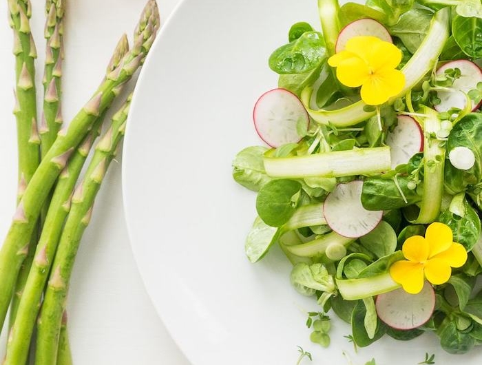 weißer teller mit salat mit grünen blättern eines feldsalats lange grüne spargel stangen zitrone gelbe blumen