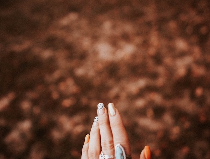 oranger nagellack eine hand einer frau mit zwei blauen großen ringen viele kleine blätter im wald