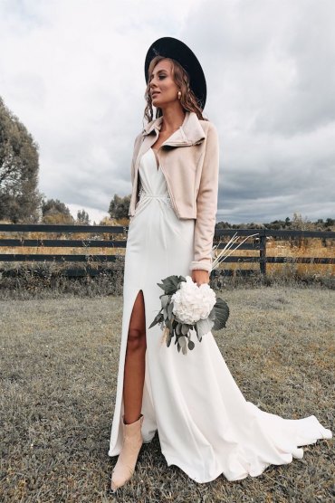 winter brautjacke beige farbe kunstpelz jacke langes weißes klied mit schlitz accessoires schwarzer hut boho brautkleid schöner strauß mit weißer blume hippie style