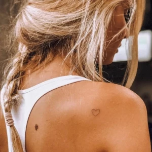 blonde frau lange haare im zopf weißes top originelle tattoo ideen minimalistisch kleines schwarzes herz tätowierung