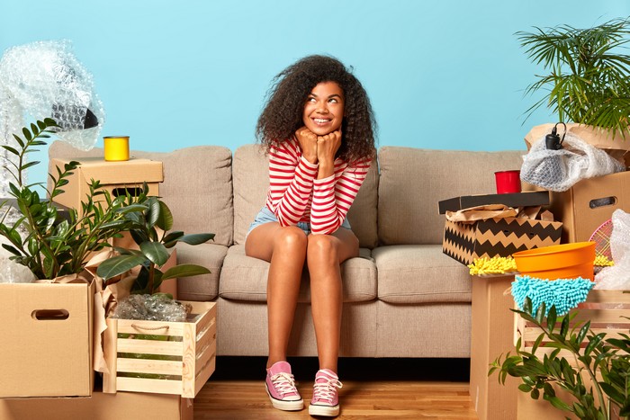 delighted female apartment owner has dreamy expression, dressed in casual clothes, sits on sofa at cozy living room, carton boxes around, thinks about good future in own rental house, looks up