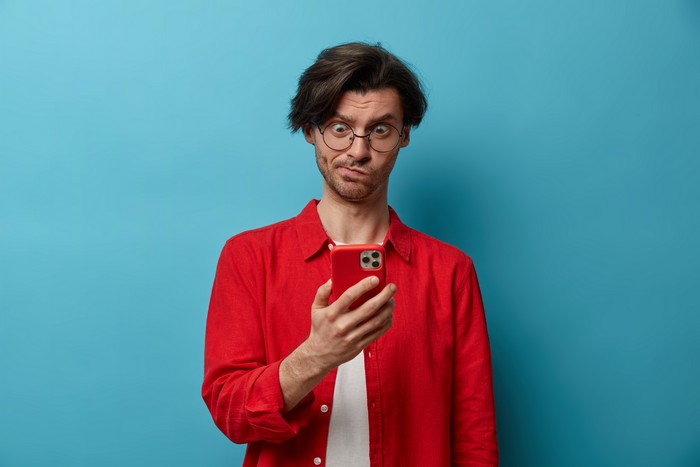 puzzled brunet youngster concentrated in smartphone display, types text message, connected to wireless internet, scrolls news, dressed in red shirt, wears optical glasses for vision correction