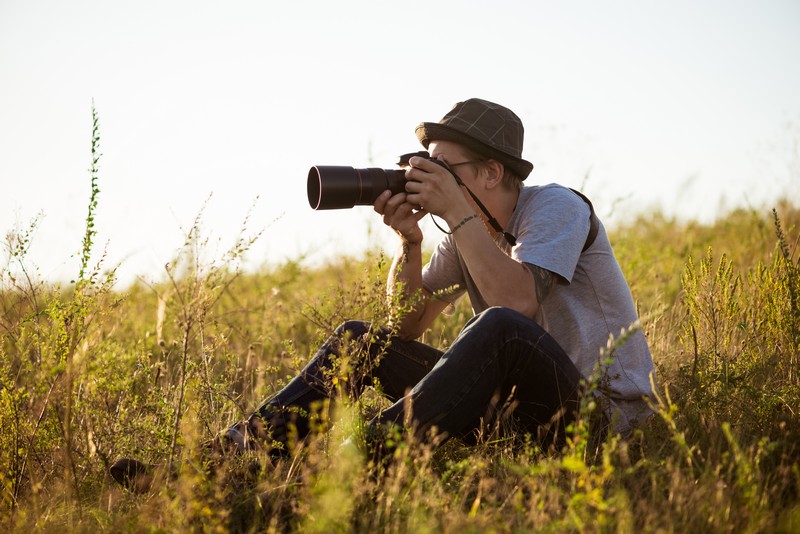 young male photographer in hat taking picture, sitting in field.