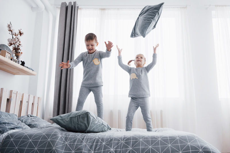 happy kids playing in white bedroom. little boy and girl, brother and sister play on the bed wearing pajamas. nightwear and bedding for baby and toddler. family at home