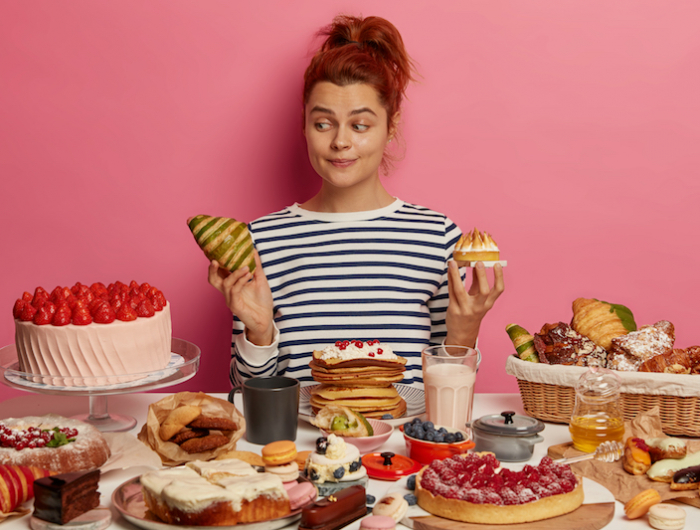 pretty redhead woman chooses between croissant and cake, sits at table overloaded with desserts, dressed in striped jumper, isolated over pink wall. unhealthy eating, harmful food and sugar obsession