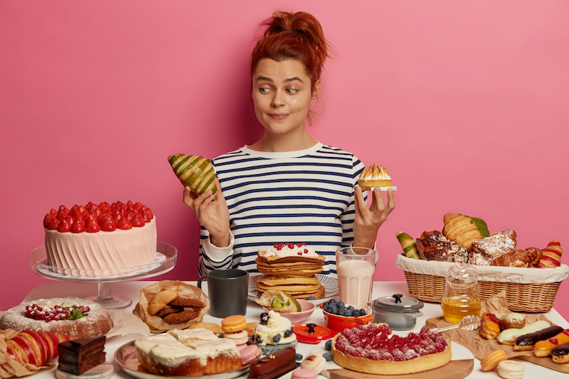 pretty redhead woman chooses between croissant and cake, sits at table overloaded with desserts, dressed in striped jumper, isolated over pink wall. unhealthy eating, harmful food and sugar obsession