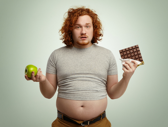 indoor shot of uncertain confused plump young male facing hard choice as he has to chose between fresh organic apple in one hand and delicious bar of chocolate in other. dilemma, diet and food