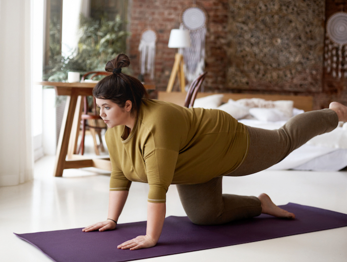 sports, activity, fitness and weight loss concept. indoor image of concentrated self determined young plus size woman in leggings and t shirt exercising on mat, lifting one leg, trying to hold balance