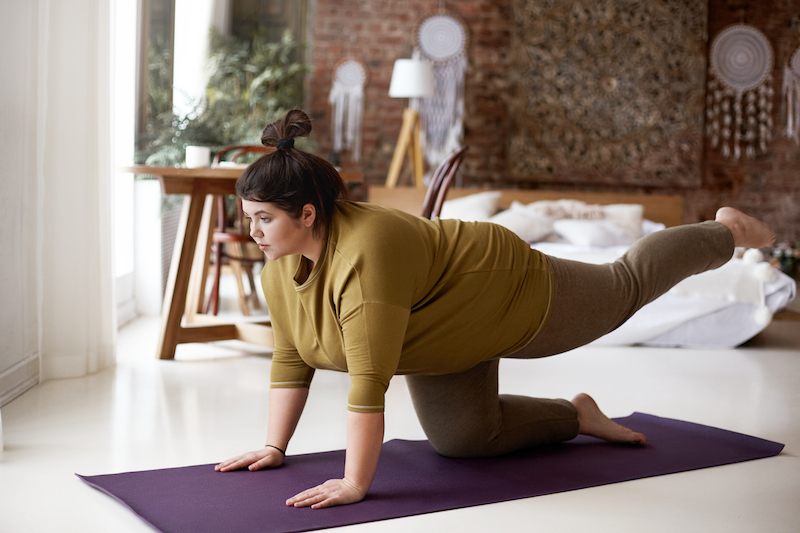 sports, activity, fitness and weight loss concept. indoor image of concentrated self determined young plus size woman in leggings and t shirt exercising on mat, lifting one leg, trying to hold balance