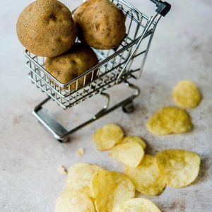 kartoffelchips, dahinter rohe kartoffeln in miniatur einkaufswagen