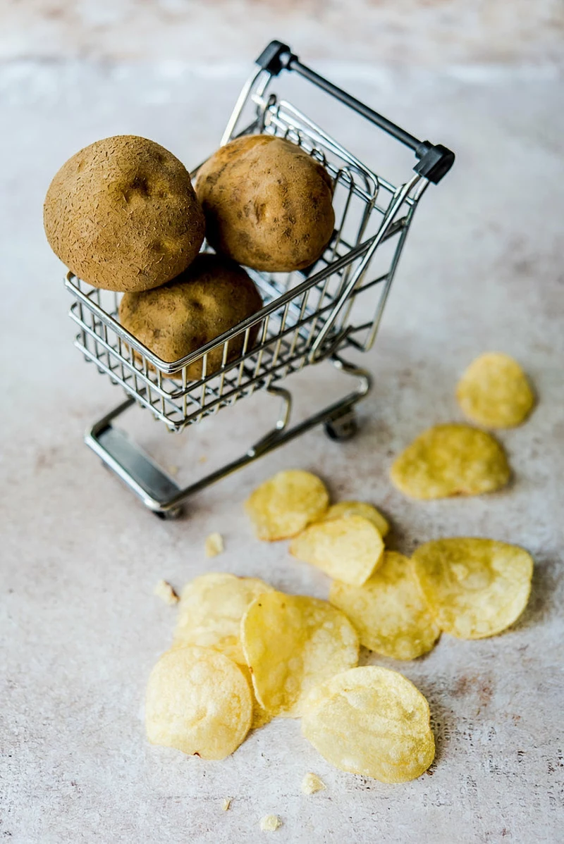 kartoffelchips, dahinter rohe kartoffeln in miniatur einkaufswagen