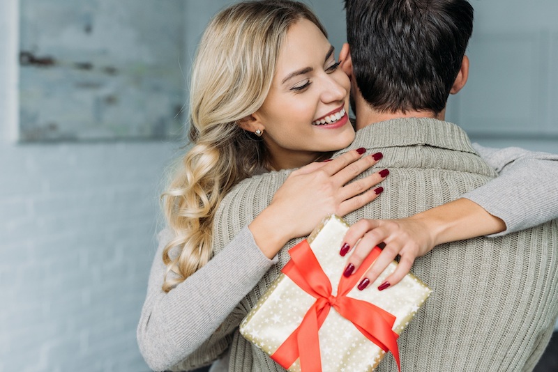 attractive young woman with gift box embracing her boyfriend at home